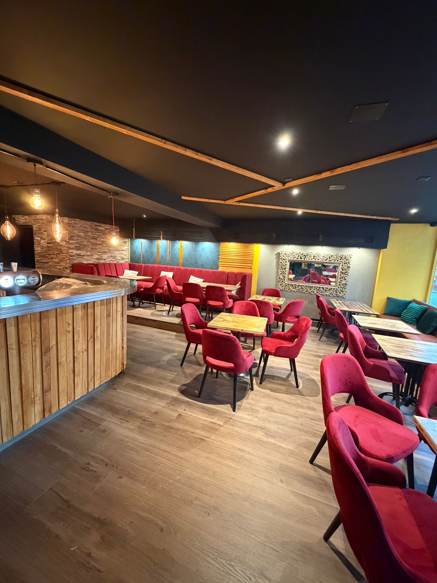 Vue large de la salle lounge d'Origen Riviera avec comptoir en bois, fauteuils rouges et lumière chaude.